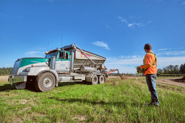 A man in an orange safety vest watches as a truck dispenses soil.