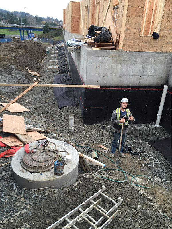 A construction site. A smiling worker stands holding a shovel and gives a thumbs up sign.